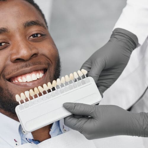 Young african man looking at camera and smiling while female dentist keeping range of fillings in clinic. Happy patient enjoying white beautiful smile while visiting dentist office. Concept of cure.