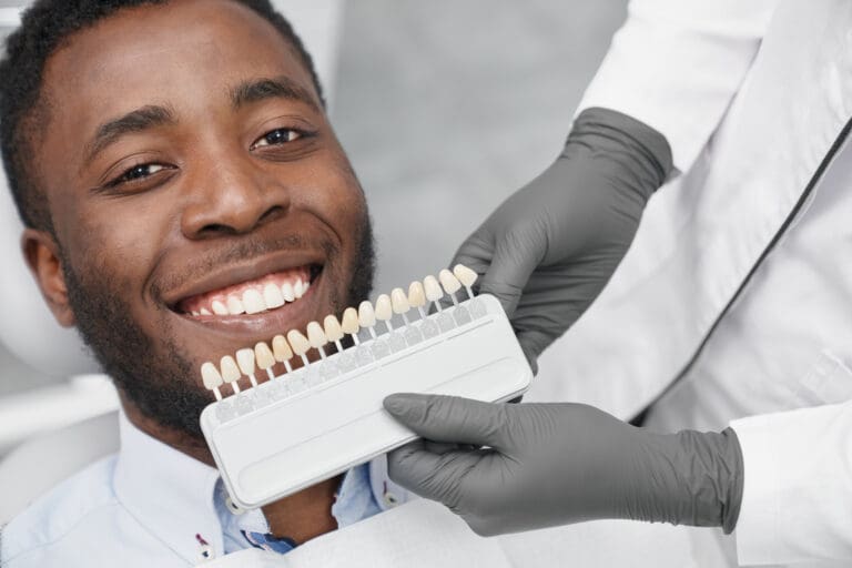 Young african man looking at camera and smiling while female dentist keeping range of fillings in clinic. Happy patient enjoying white beautiful smile while visiting dentist office. Concept of cure.