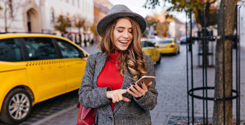 woman texting message while walking on the street. Lady in coat looking at phone screen, standing on avenue with taxi on background.